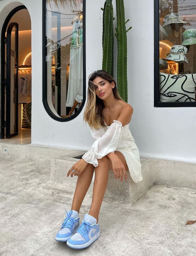 A woman sits on a concrete ledge outside a white building in a white off-the-shoulder dress and sneakers, with a cactus behind her.
