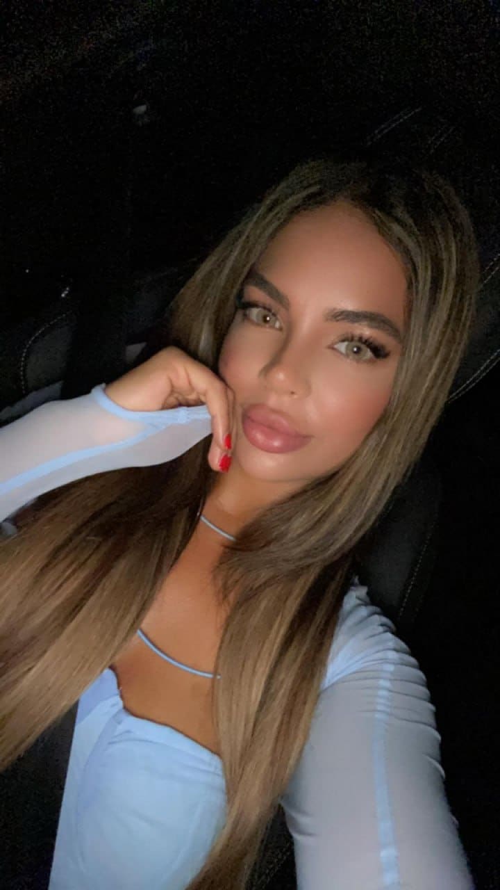 A woman with straight brown hair sits in a dark car, wearing a light blue top with sheer overlay and a choker, resting her chin on her red-nailed hand.
