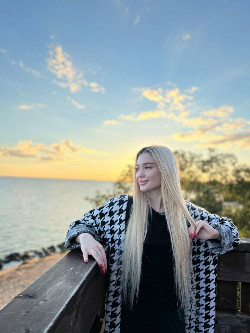 Evita stands outdoors at sunset, leaning on a wooden railing in a black top and houndstooth coat, with water and trees behind her.