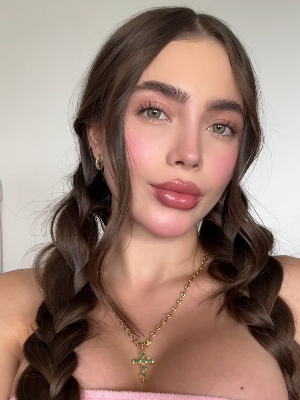 Close-up of a woman with braided brown hair, wearing a pink strapless top and gold cross necklace against a light wall.