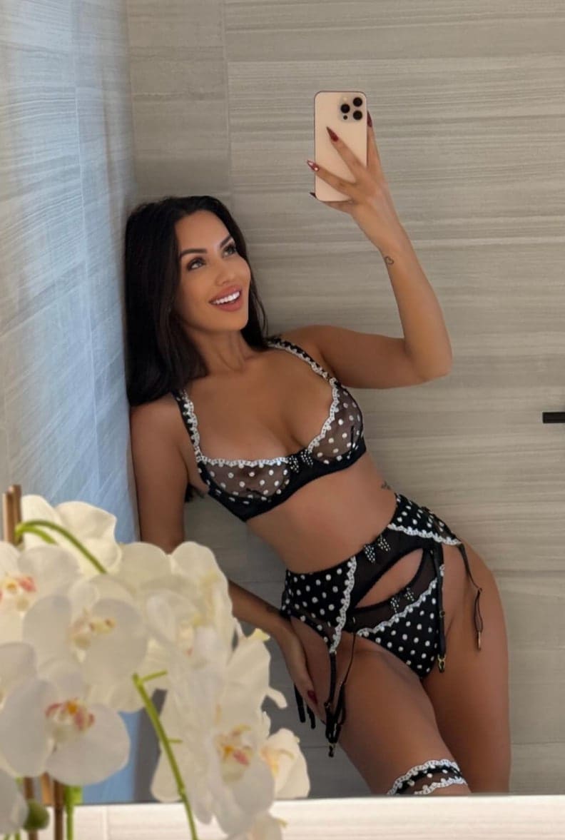 Mirror selfie of a woman in a black and white polka-dot lace lingerie set, standing in a tiled room with white orchids in the foreground.