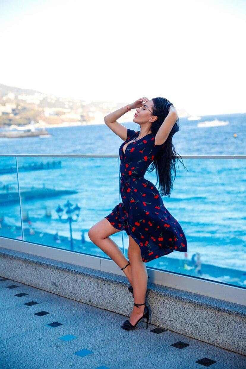 A woman stands on a tiled terrace in a floral dress and heels, hand to forehead, near a pool and sea.