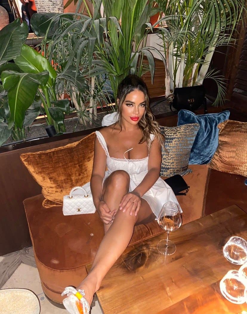 A woman sits on a brown velvet sofa in a short white dress with thin straps, legs crossed and hand on knee, surrounded by potted plants and pillows indoors.