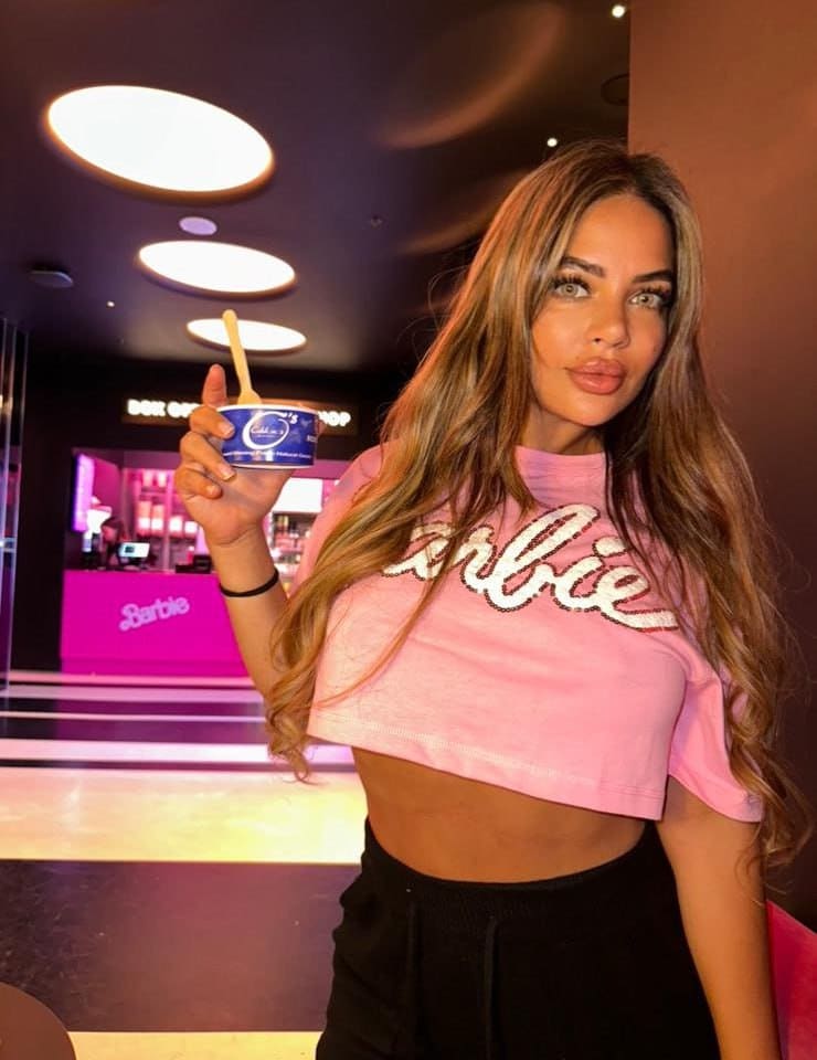 A woman stands indoors holding a blue cup, wearing a pink cropped t-shirt with white lettering and black pants, near a pink counter and round ceiling lights.