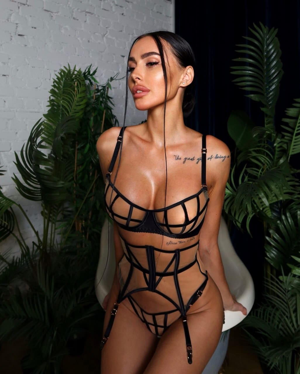 A woman sits on a white chair before a brick wall and plants, wearing a black strappy lingerie set.