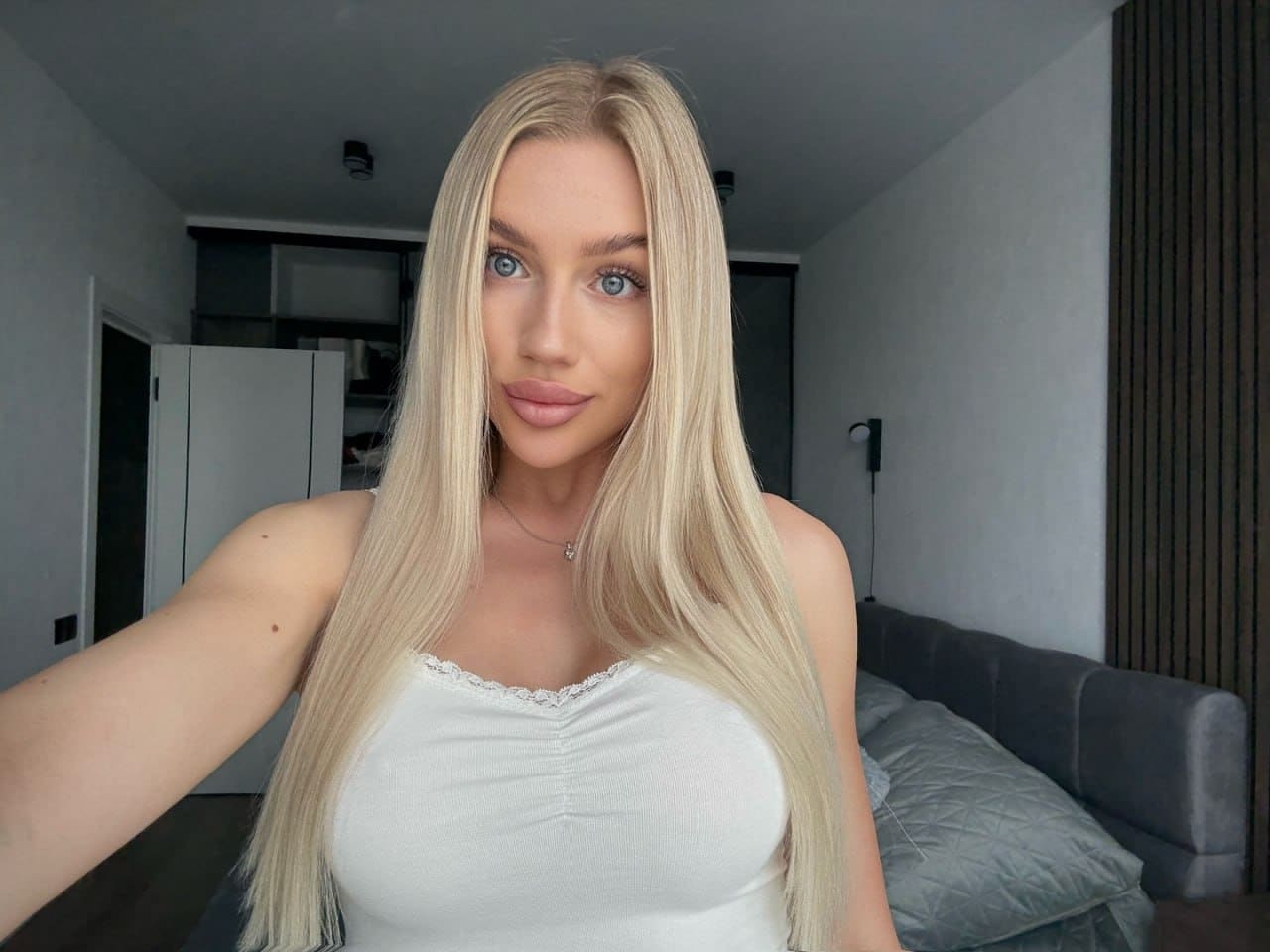 Arianna takes a bedroom selfie in a white lace-trimmed top, with a grey sofa and wardrobe in the background.