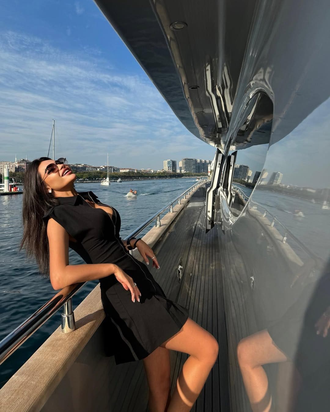 Leaning on a boat railing, she wears a short black dress and sunglasses, with a waterfront cityscape in background.