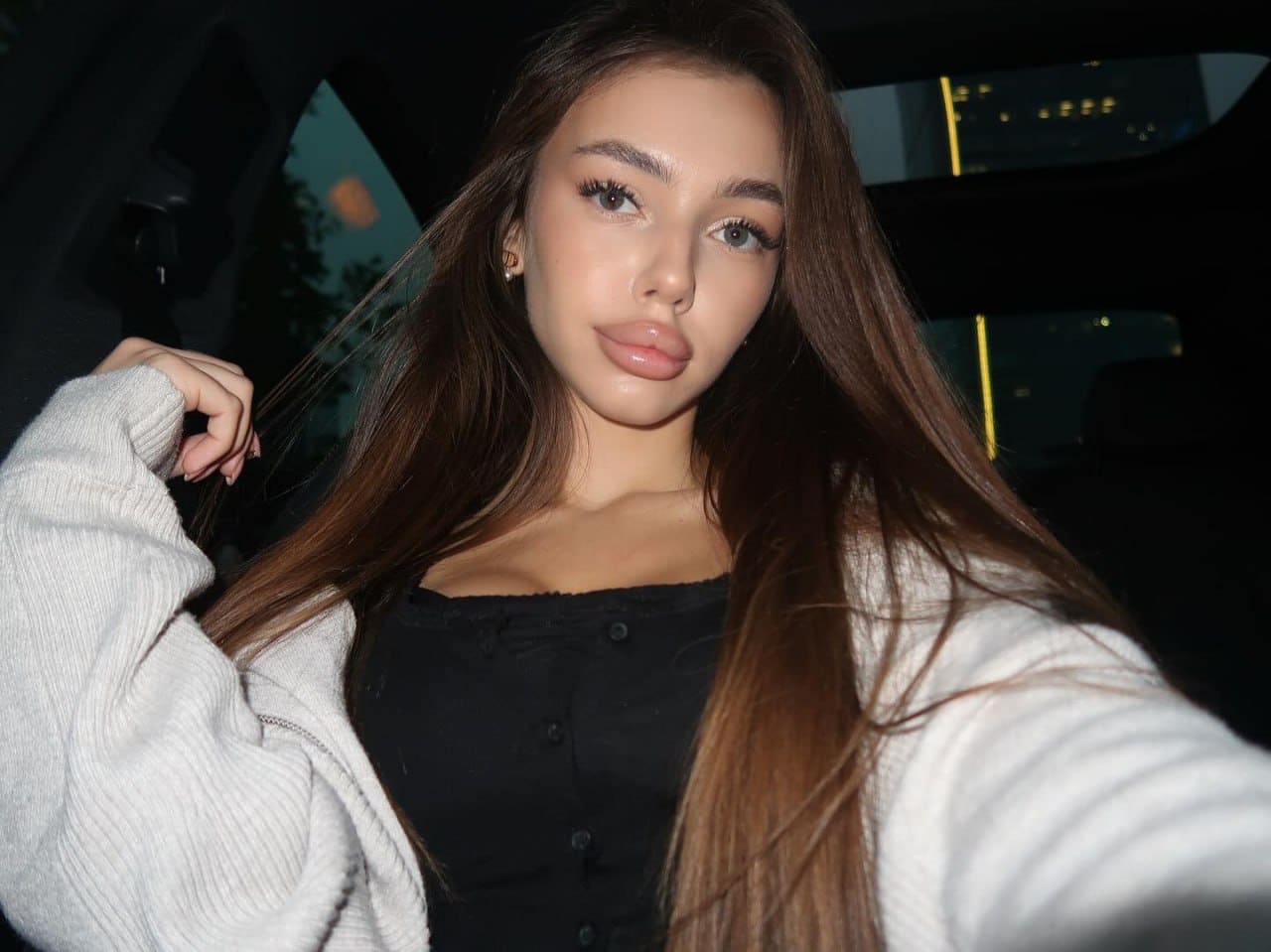 A woman takes a night selfie inside a car, wearing a black button-up top under an open white cardigan, with one hand raised near her hair.