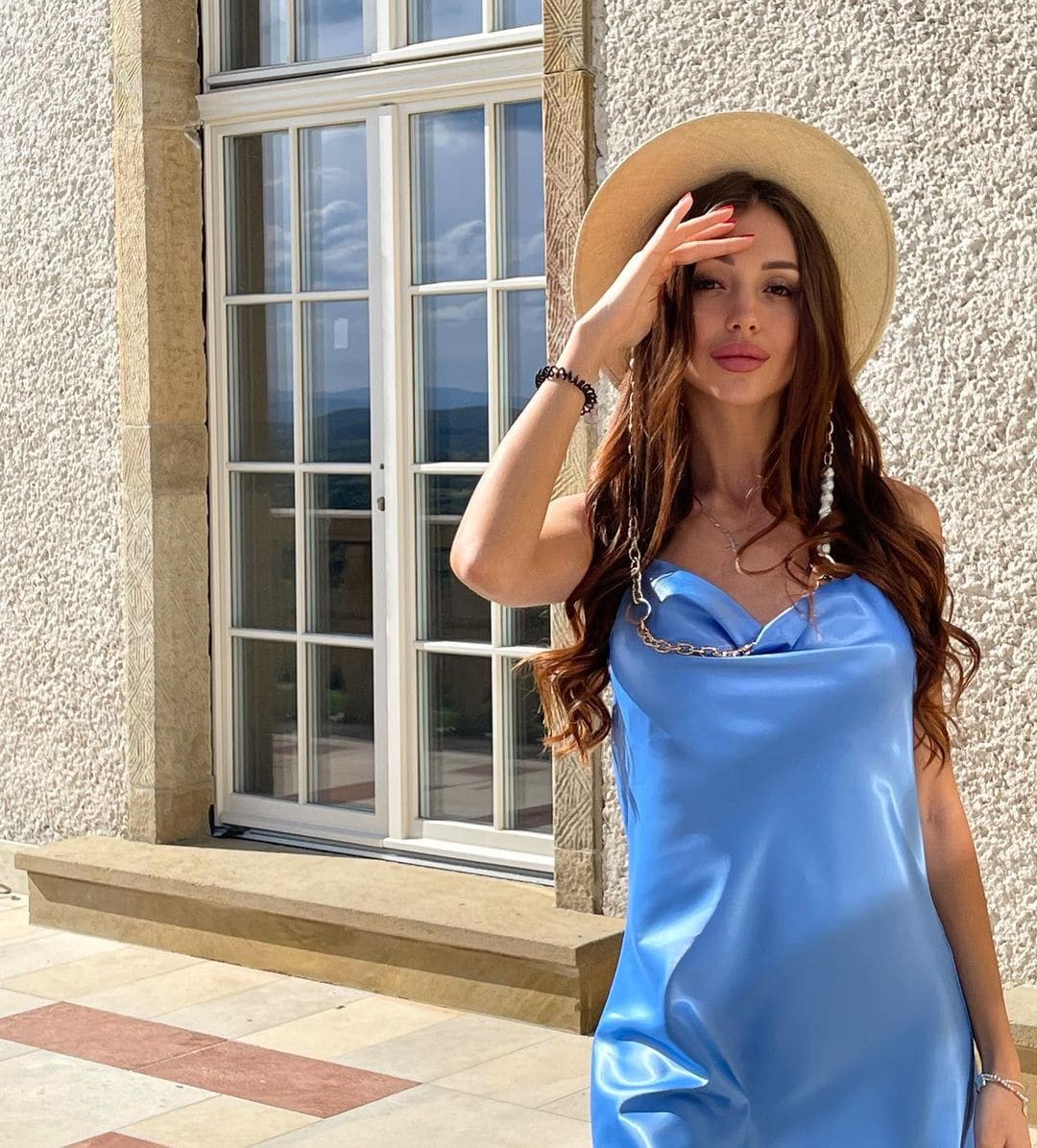 Dalfina wears a straw hat and light blue satin dress outdoors, shading her eyes in front of a stone building with a large window.