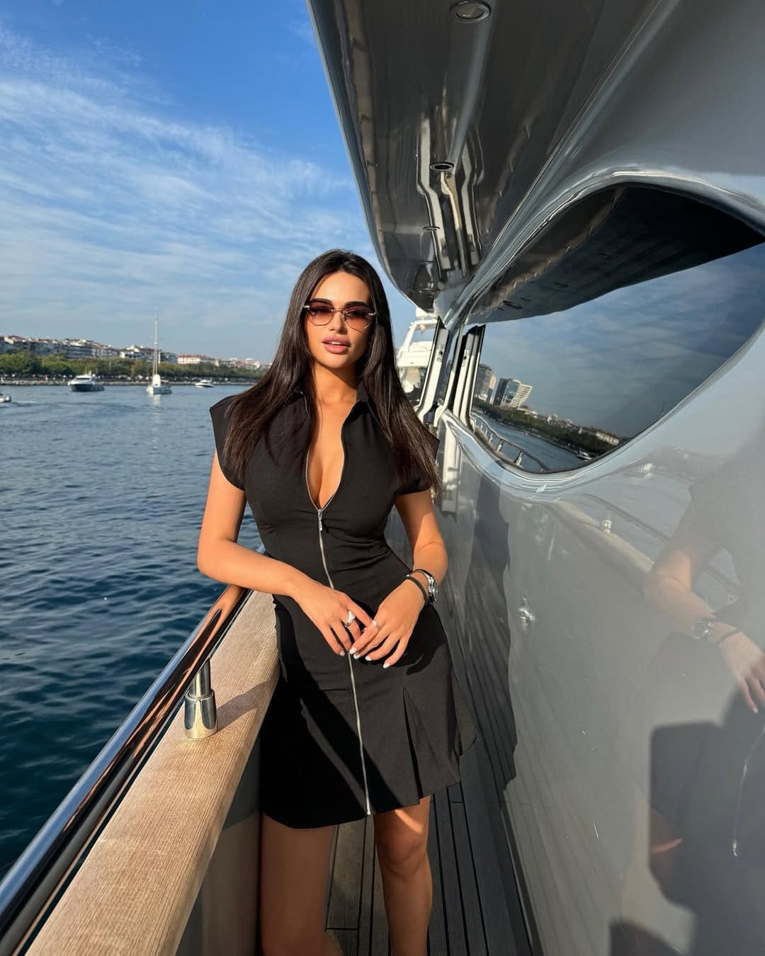 On a boat deck, she wears a black zip-front dress and sunglasses, hands near the zipper, water and shore behind.