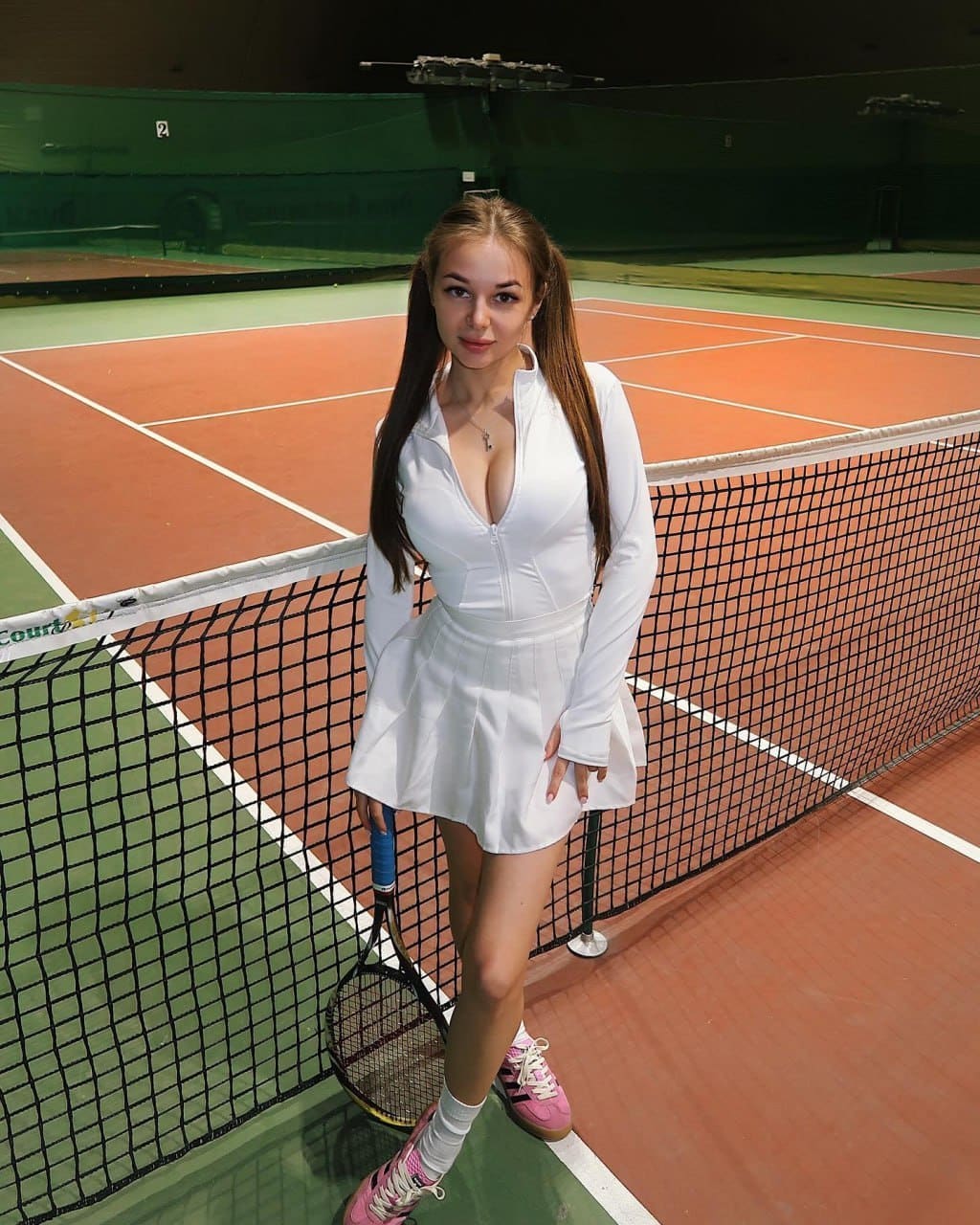 A woman with pigtails holds a racket on a clay court, dressed in a white zip-up top, pleated skirt, white socks, and pink sneakers.