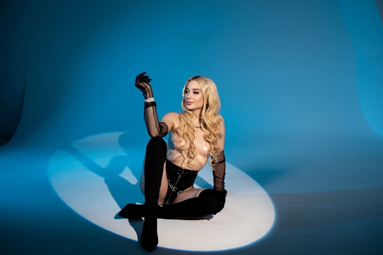 A woman sits on a white platform against a blue backdrop, wearing a black corset, sheer sleeves, and thigh-high boots, with one hand raised.