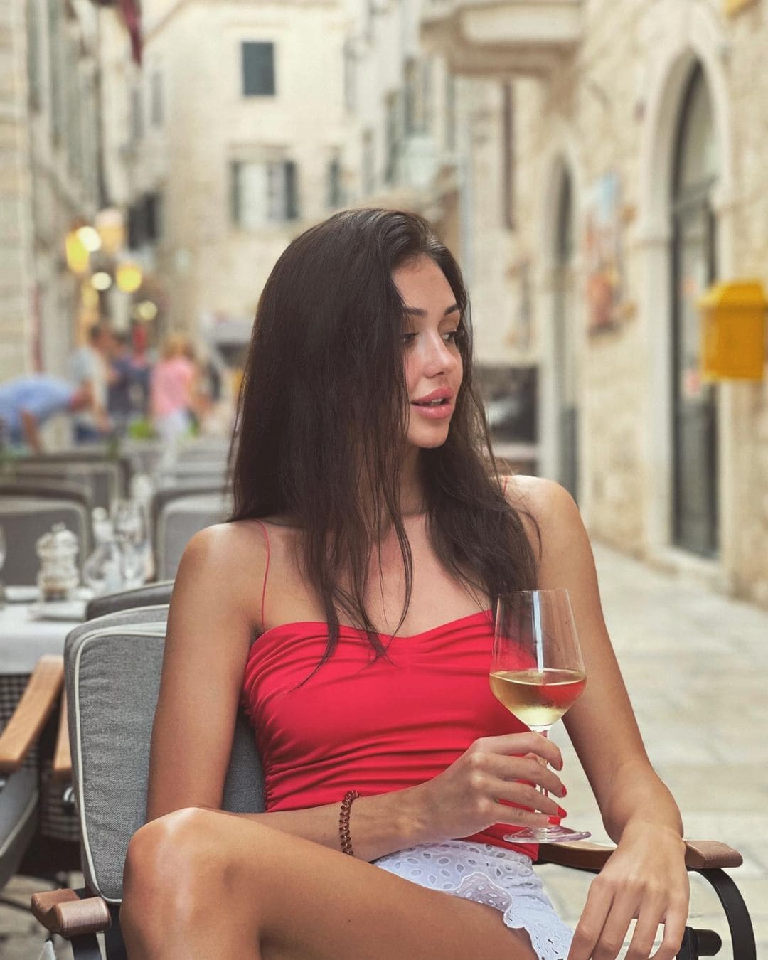 A woman in a red top and white lace skirt sits at an outdoor café, holding white wine and looking sideways on a stone-paved street.