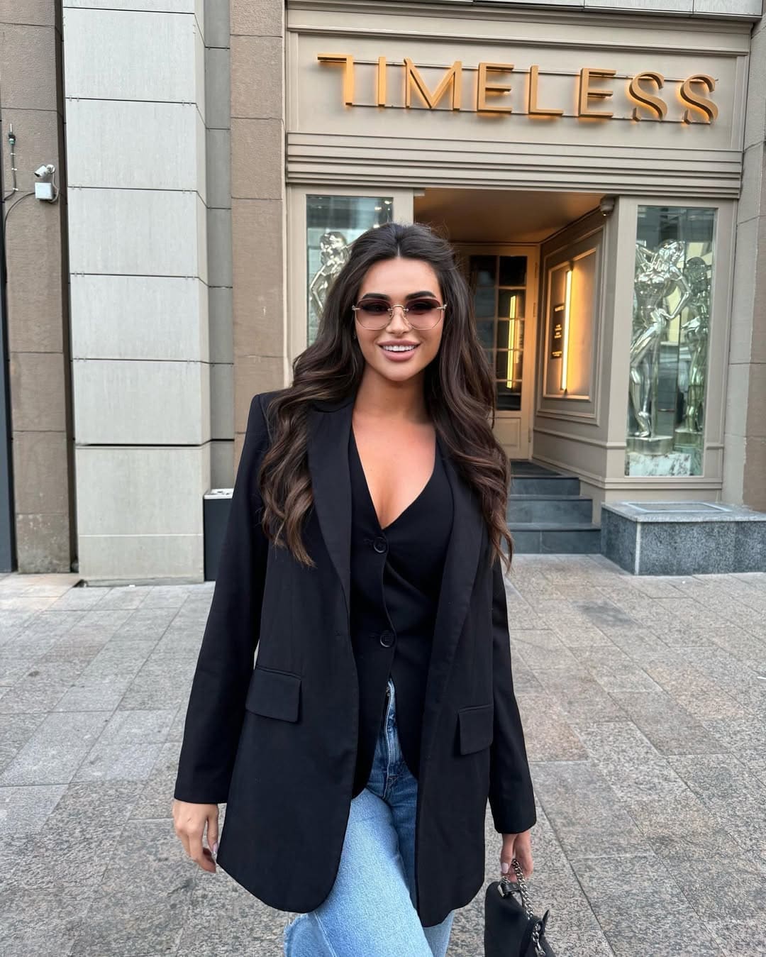 Helena stands on a sidewalk in a black blazer, V-neck top, and light blue jeans, holding a small black handbag.
