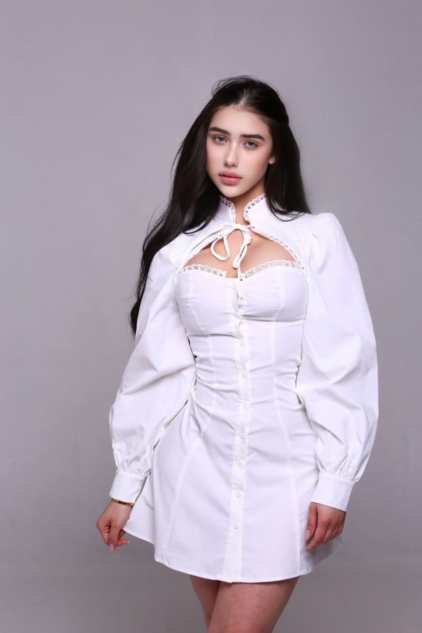 Wearing a white long-sleeved dress with a chest cutout, she stands facing forward against a gray studio backdrop.