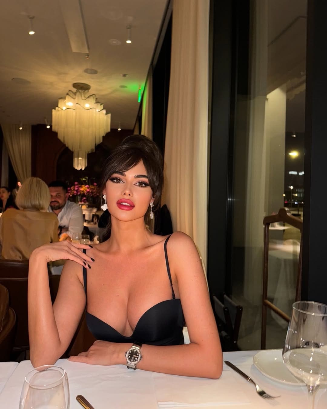 Seated at a restaurant table, she wears a black spaghetti-strap top, wristwatch, and earrings, hand on shoulder.