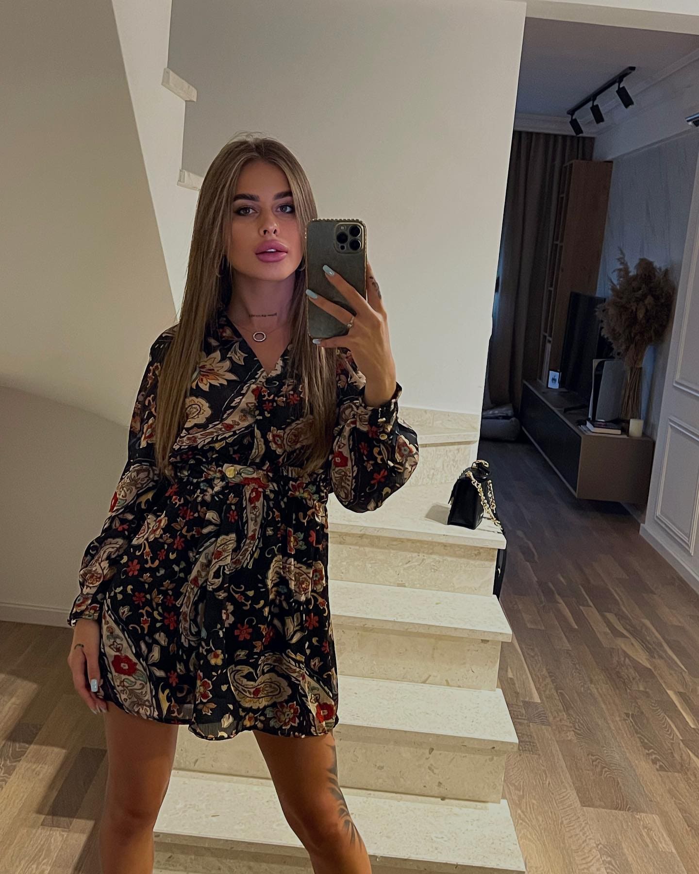 A woman stands on stone steps in a black floral dress, taking a mirror selfie with a phone, while a small black handbag rests on the step beside her.