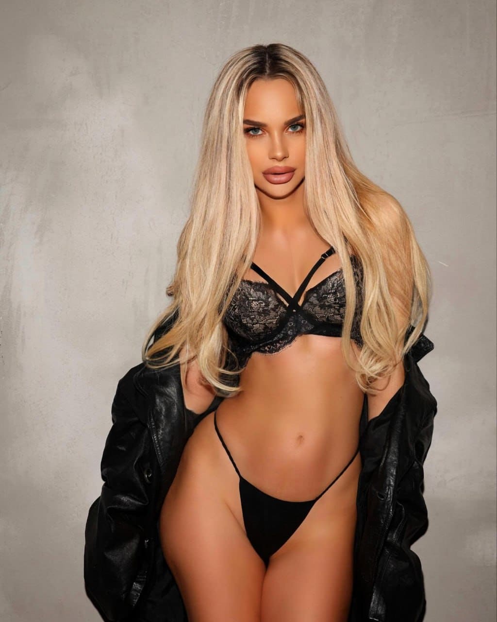 Miminel stands facing the camera in black lace lingerie and an open leather jacket against a plain grey wall.