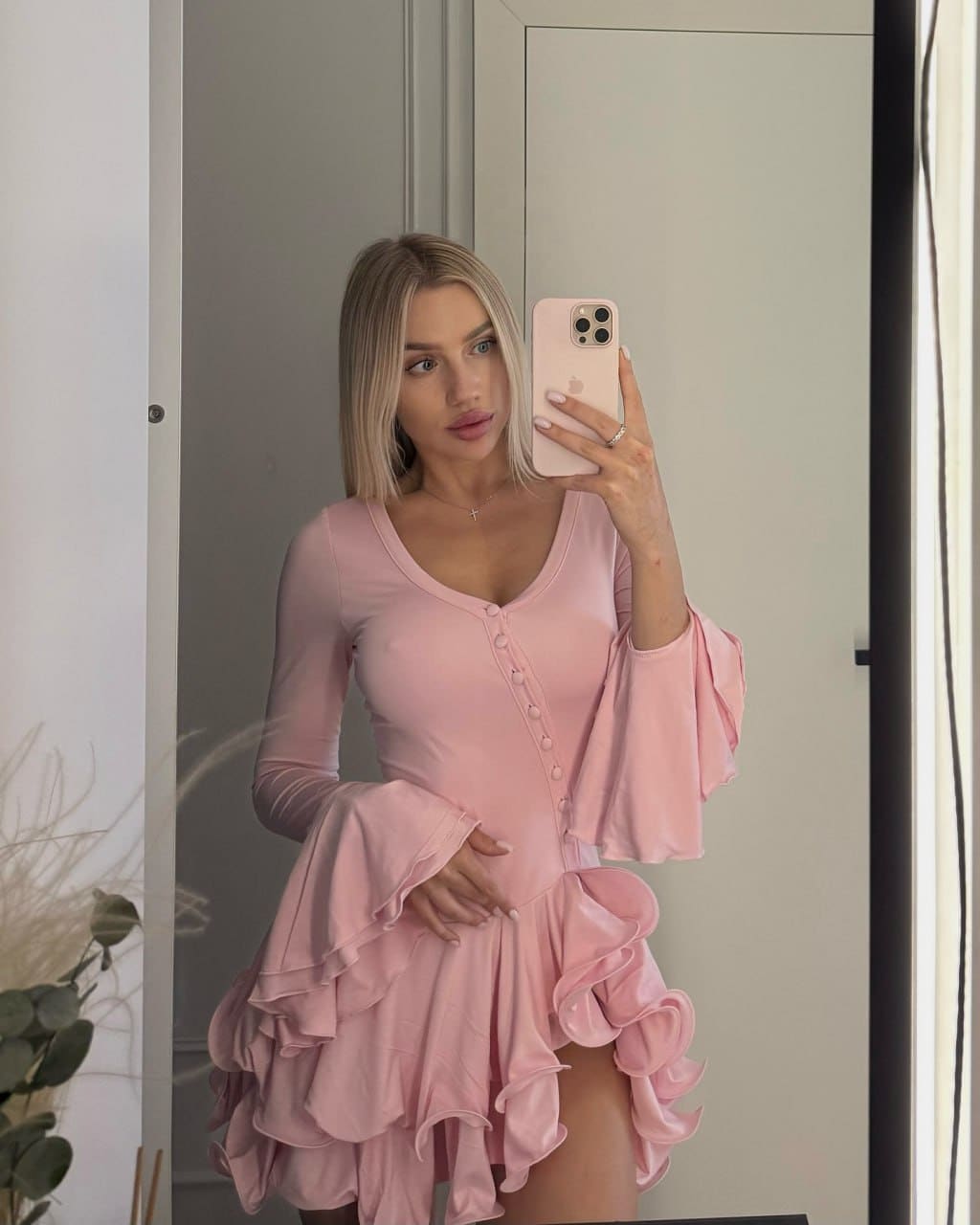 A blonde woman takes a mirror selfie in a pink ruffled dress, standing by a white door with a plant nearby.