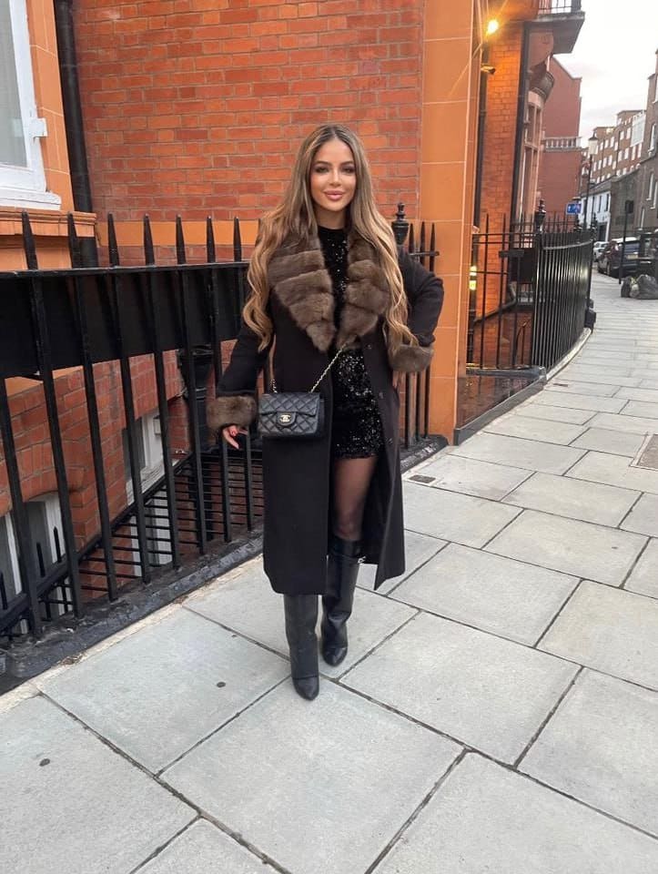 A woman stands on a sidewalk before a brick building, wearing a black sequined dress under a fur-collared coat, black tights, knee-high boots, and a crossbody bag.
