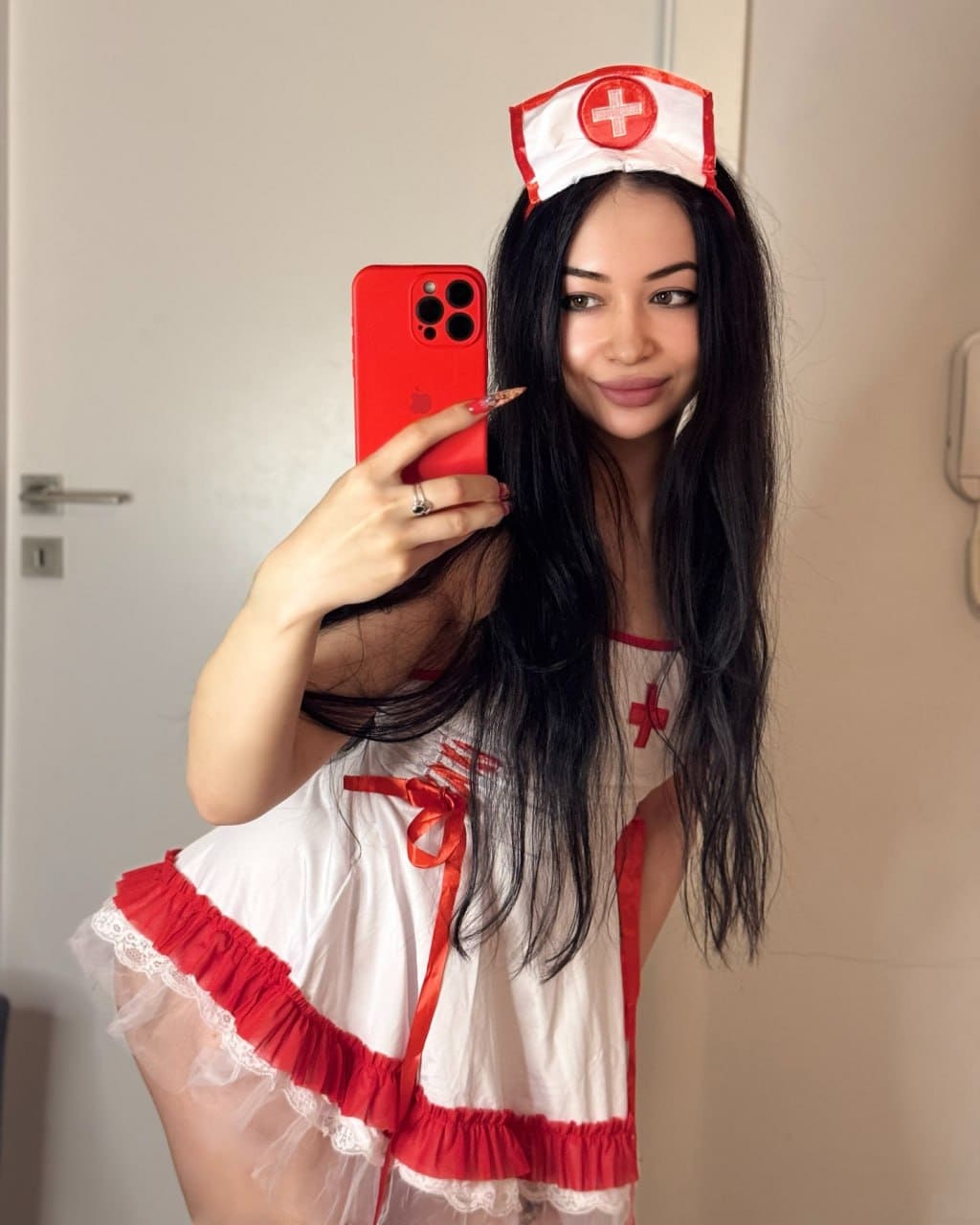 Mirror selfie of a woman in a red and white nurse costume holding a red smartphone against a plain wall.