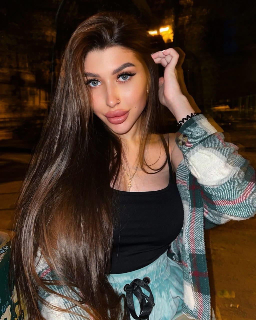 Aureline poses outdoors at night in a black tank top, open plaid flannel, and light blue drawstring pants, one hand raised to her long brown hair.