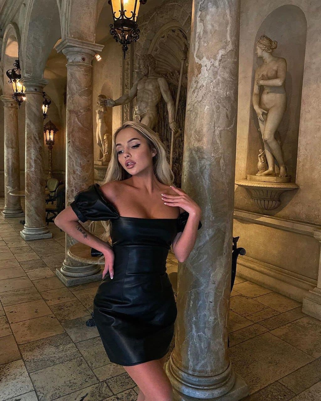 A blonde woman in a black leather dress poses with a hand on her hip in a marble hall with statues and lanterns.