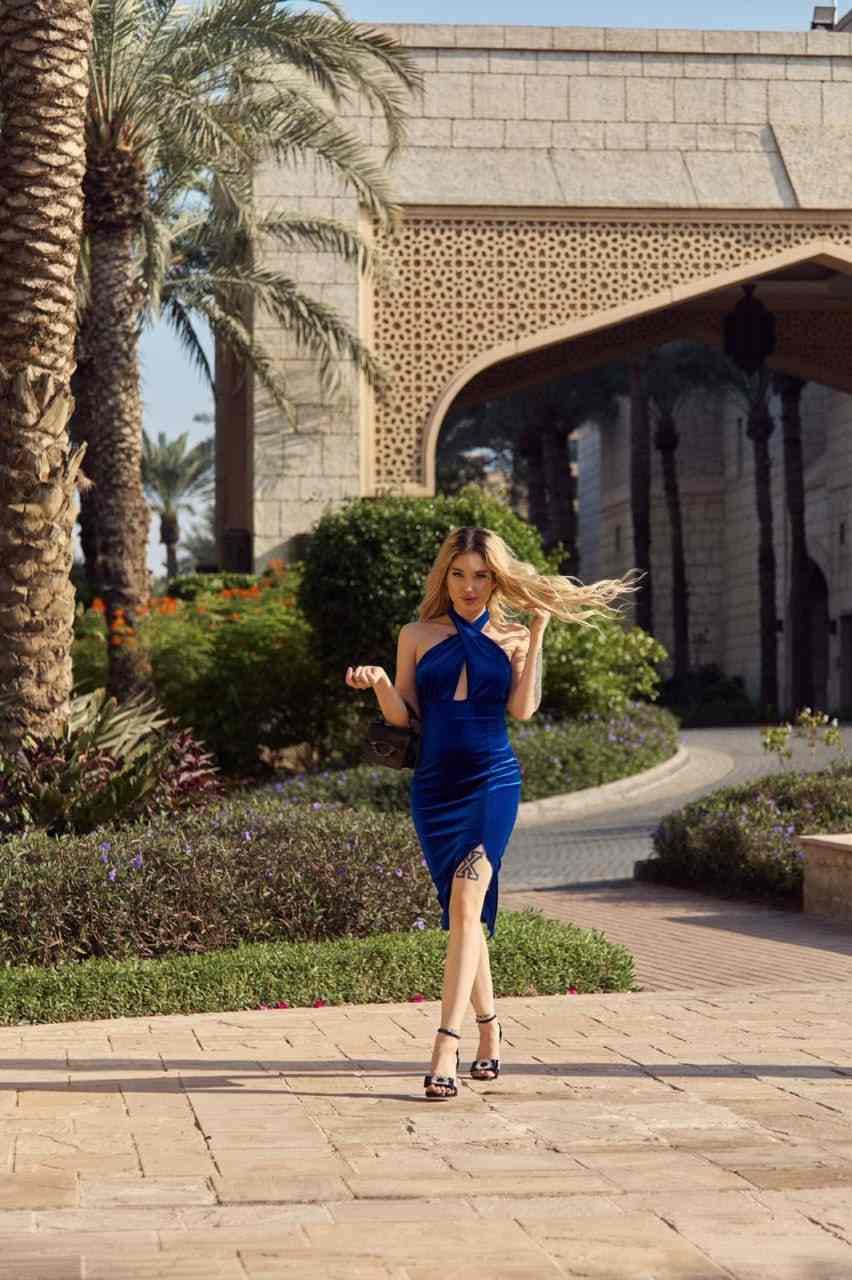 A woman stands on a stone walkway in a blue halter dress with a slit, holding a dark handbag, with a stone building and palm trees behind.