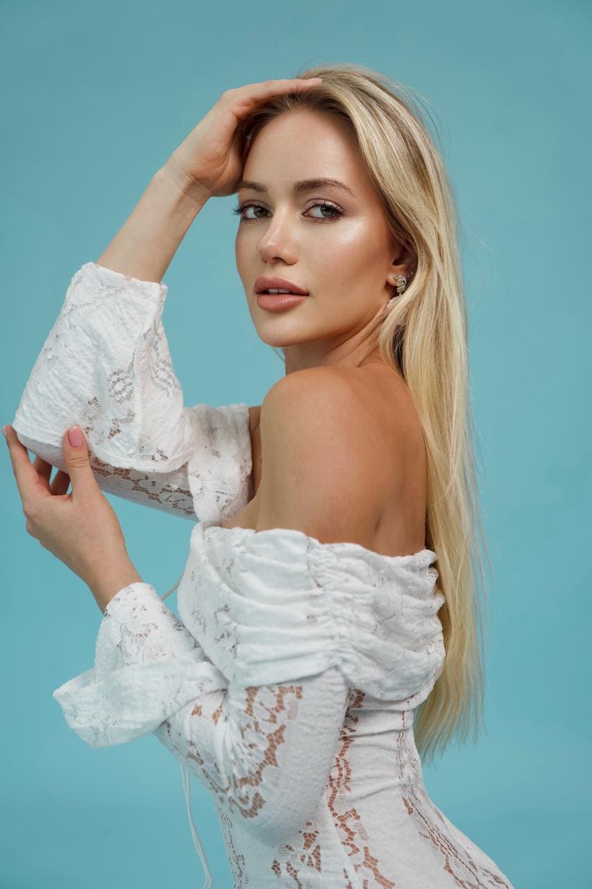 Inessa wears a white off-the-shoulder lace dress with bell sleeves, one hand raised to her head against a light blue background.