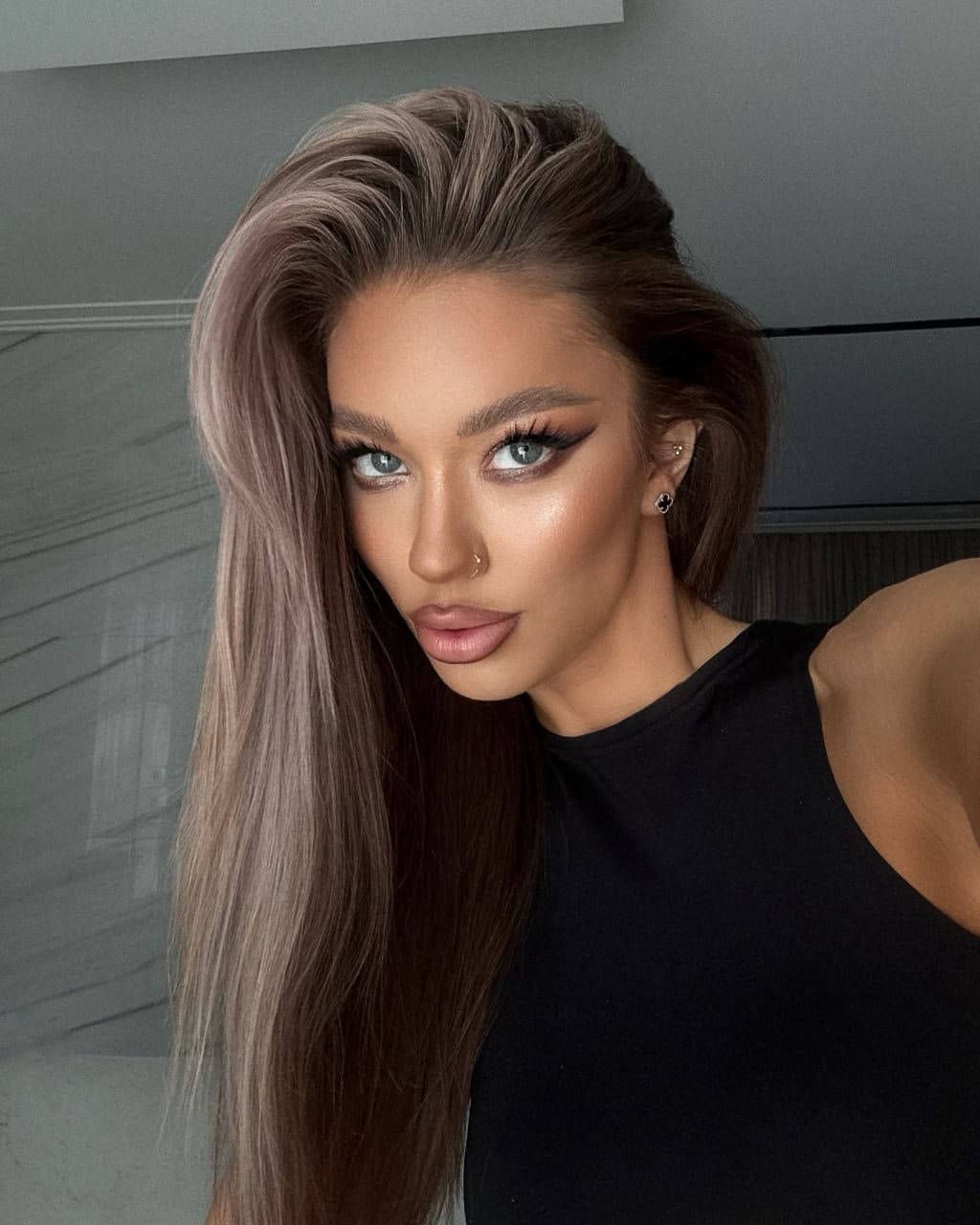 Amina in a close-up selfie with long brown hair, wearing a black sleeveless top and nose ring against a gray wall.