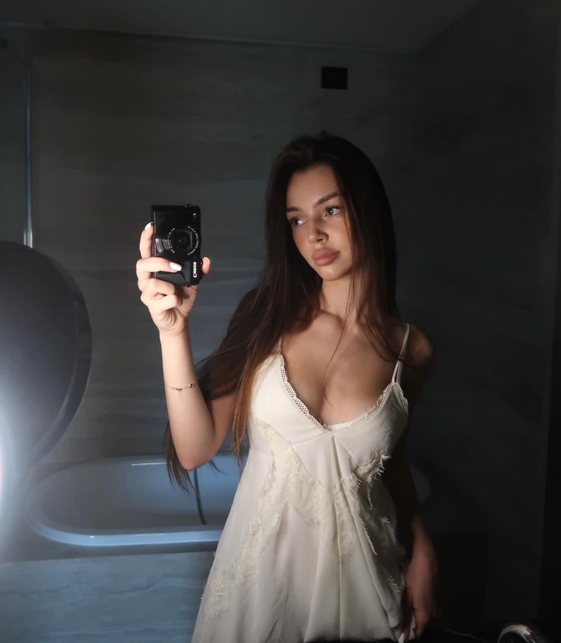 A woman takes a mirror selfie in a dim bathroom, wearing a white lace-trimmed slip dress and holding a black compact camera, with a bathtub behind.