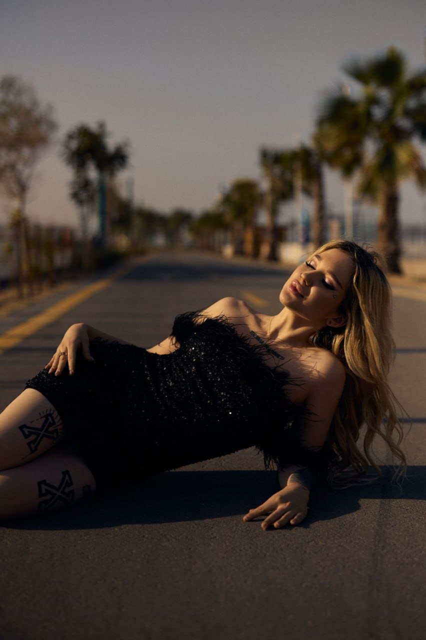 A woman lies on an asphalt road in a short black sequined dress, displaying geometric thigh tattoos, framed by palm trees under a hazy sky.