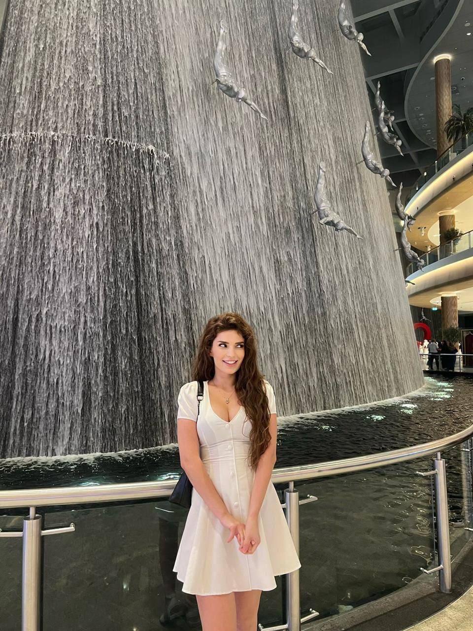 Standing before an indoor waterfall, she wears a short-sleeved white dress with a V-neck and black shoulder strap, hands clasped in front.