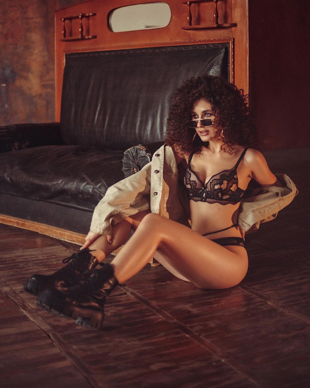 A woman sits on a wooden floor in black lace lingerie, an open jacket, and platform boots, wearing sunglasses.