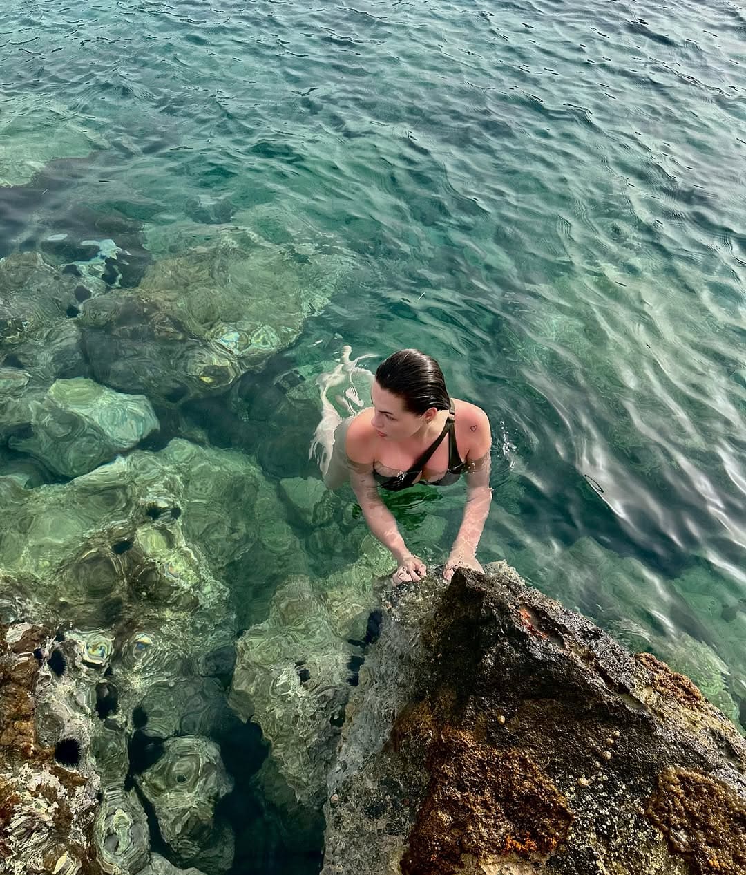 A woman with wet hair stands in clear turquoise water wearing a black bikini top, holding onto a rocky outcrop in the foreground.