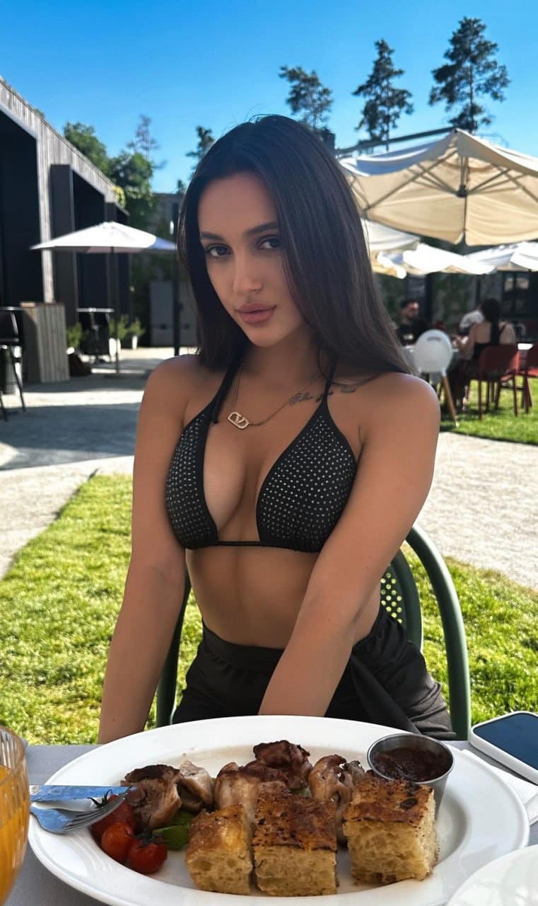 Vivien sits at an outdoor table in a black studded bikini top and dark bottoms, with a plate of food and umbrellas in the background.