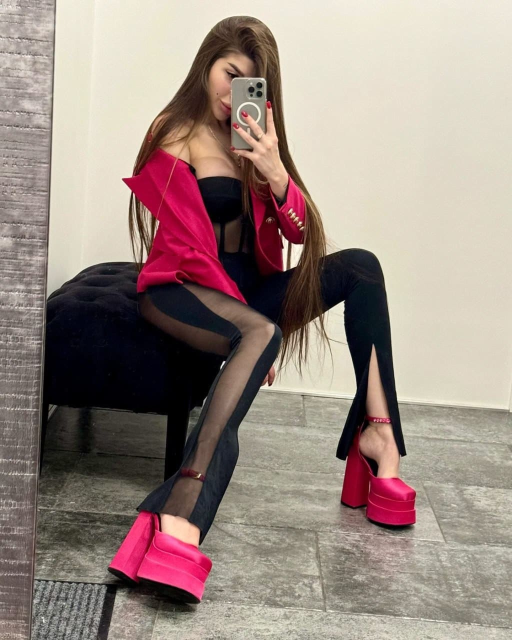 A woman sits on a black tufted chair in a grey-tiled room, wearing a black bustier, pink blazer, and platform heels, holding a phone to her face.