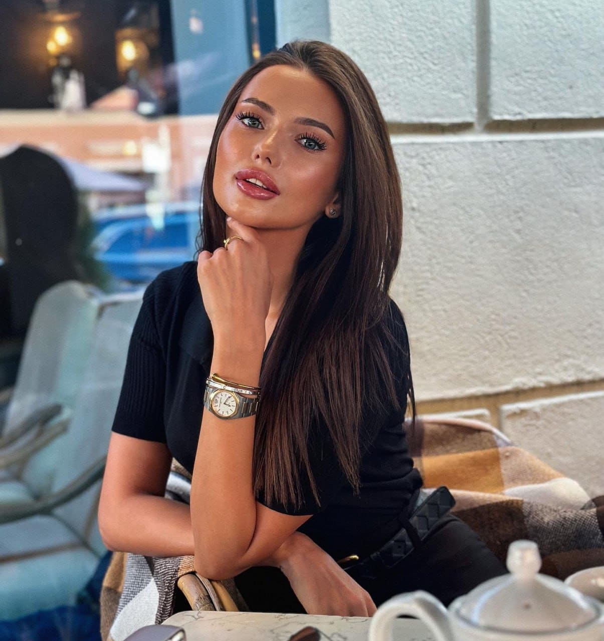 Tatyana sits at a table in a black short-sleeved top, resting her chin on her hand, with a teapot and plaid blanket in the foreground.