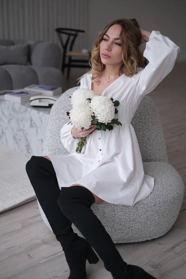 A woman sits in a white button-up dress and black boots, holding white flowers and resting a hand in her hair.
