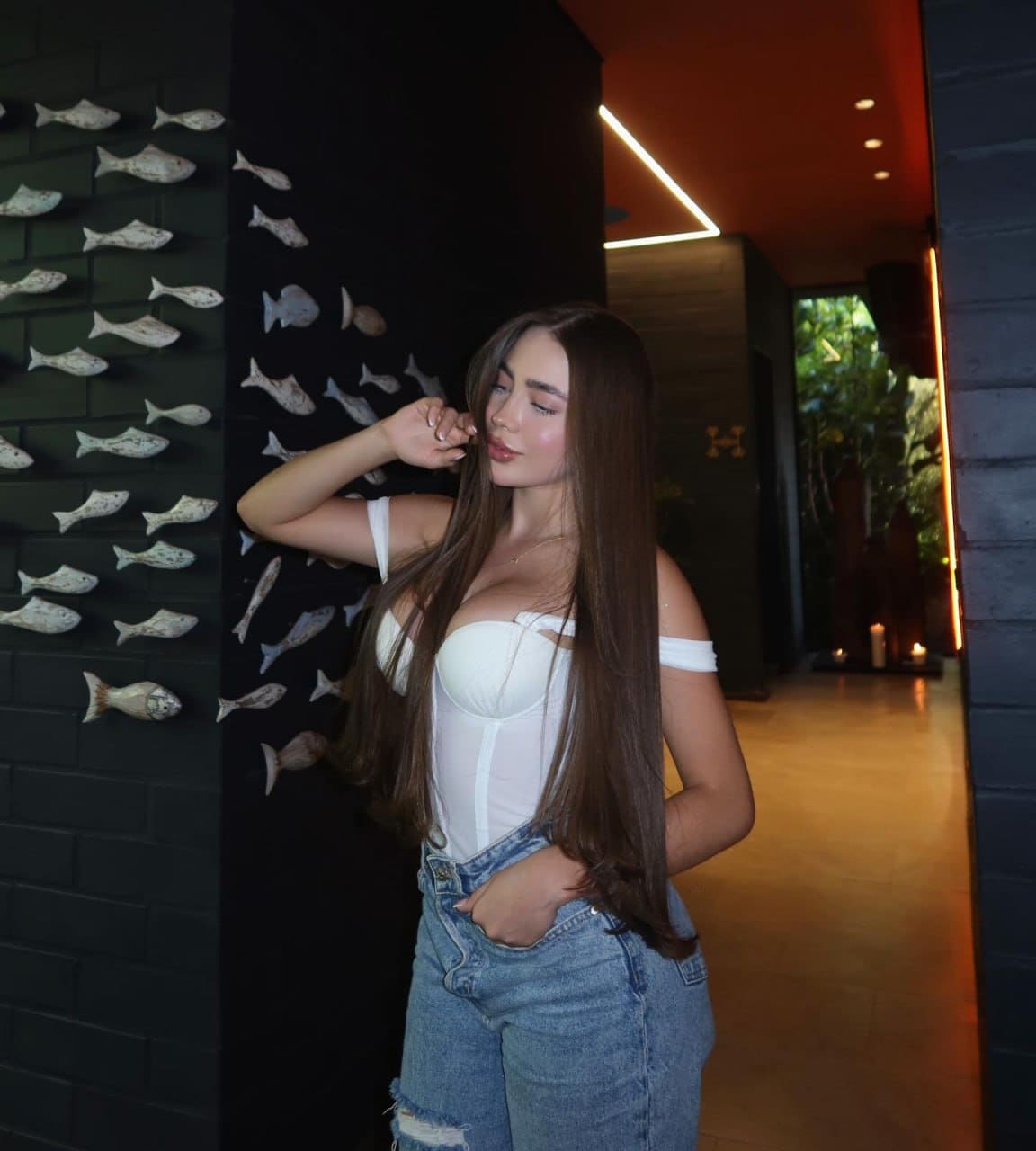Woman standing in a hallway with fish ornaments, wearing a white off-shoulder top and blue jeans, hand near face.