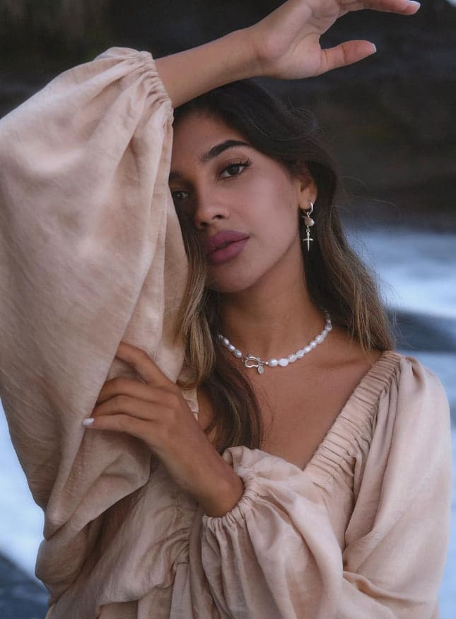 Sabrina poses outdoors in a beige V-neck blouse and pearls, holding a sleeve near her face against a blurred rocky water background.
