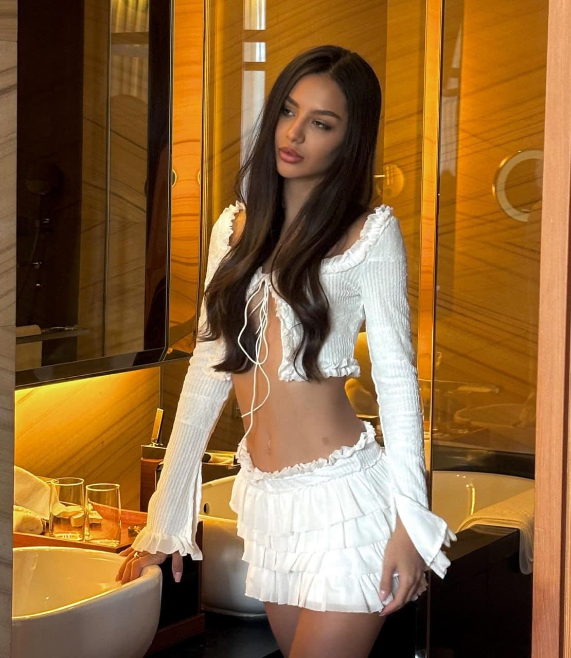 Woman stands in a bathroom wearing a white ruffled crop top and mini skirt, resting her left hand on the edge of a white sink.