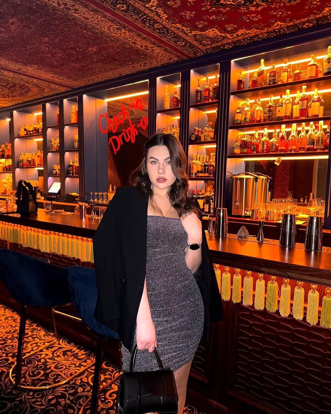 A woman stands in a bar wearing a strapless silver sequined mini dress with a high slit and a black blazer draped over one shoulder.