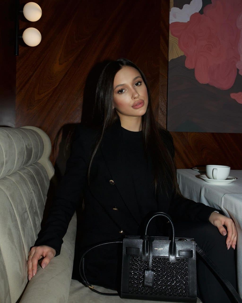 A woman in a black blazer and top sits on a beige sofa with a black handbag on her lap, in a wood-paneled room.