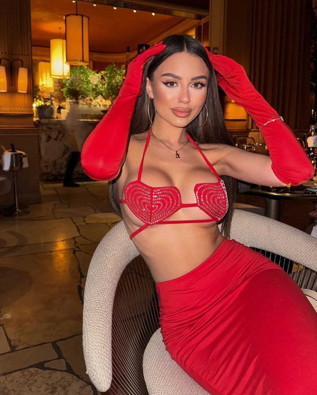 A woman sits in a woven chair wearing a red beaded halter top, matching skirt, and long red gloves, hands raised to her head.