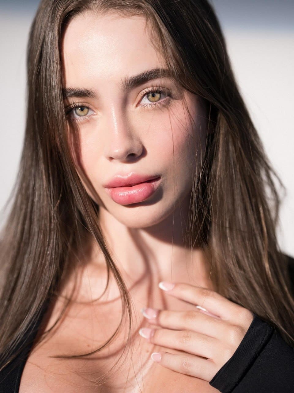 Juliette with long brown hair in a black low-neck top, hand on chest, against a plain light background.