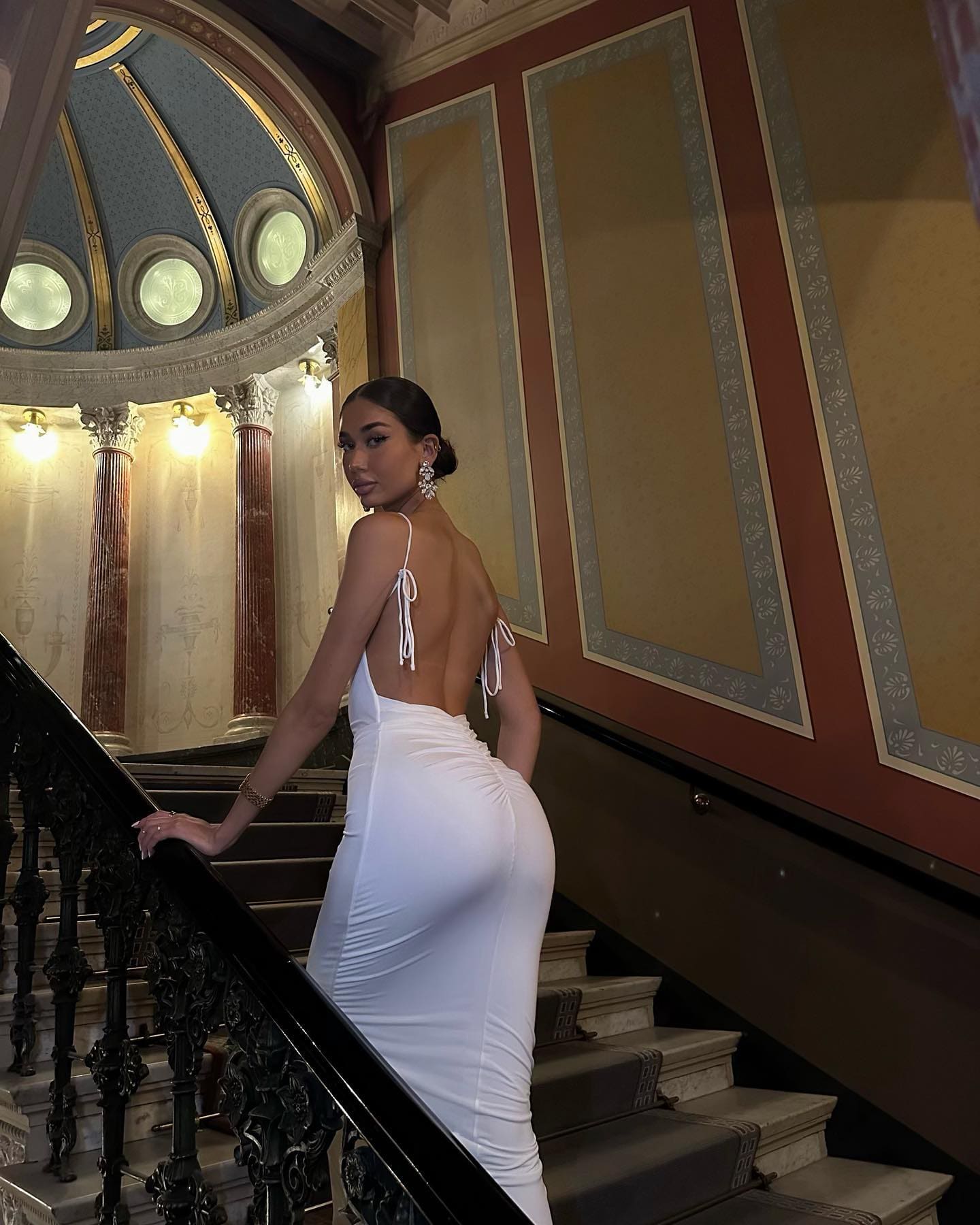 A woman in a white backless dress rests a hand on an ornate railing on a grand staircase, looking over her shoulder.