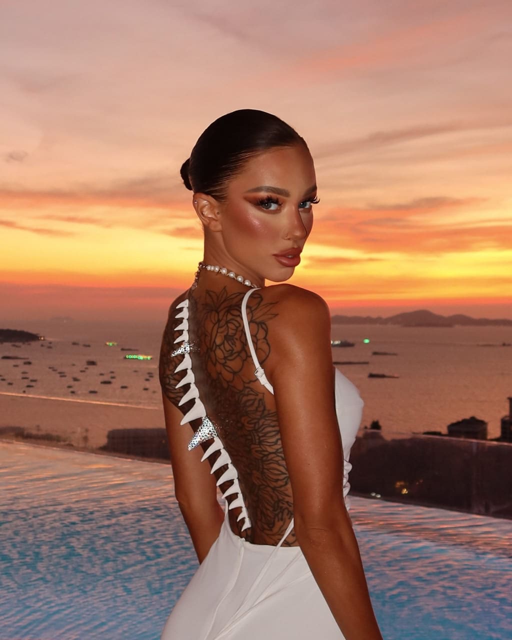 Woman in a white backless dress and pearl necklace by an infinity pool at sunset, showing a floral back tattoo.