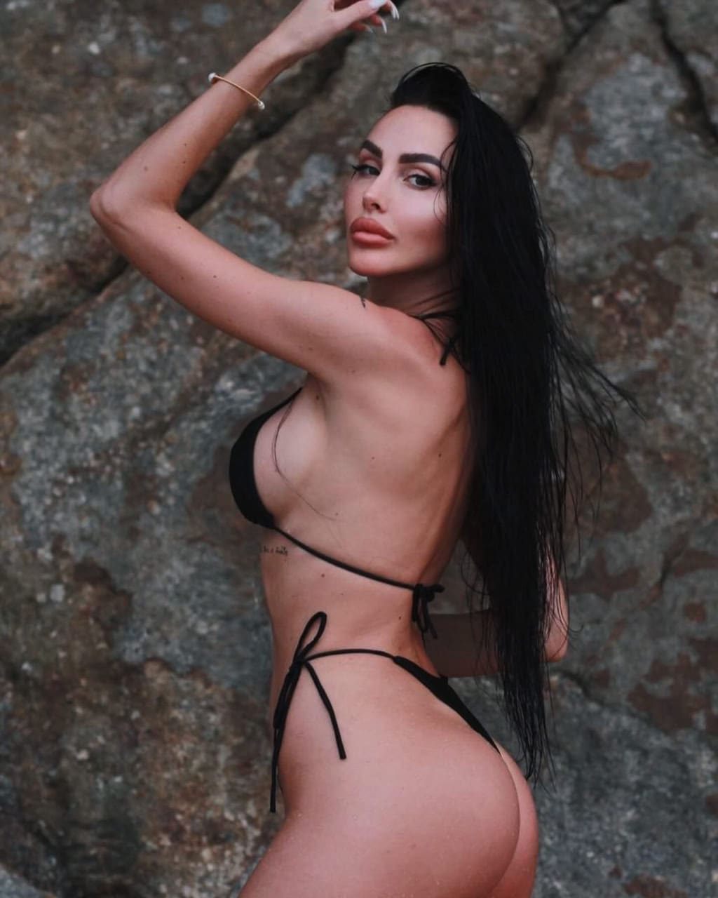 A woman poses in a black string bikini before a rocky surface, with one arm raised above her head.