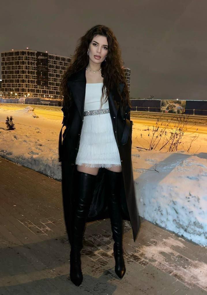At night on a snowy walkway, she wears a white fringed mini dress, black coat, and thigh-high boots, with a lit building in the background.